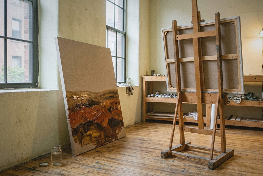A Brooklyn atelier in late morning light: a finished oil painting leaning face out against the north wall with wet impasto on its lower third, and a raw linen canvas turned to face the wall on the easel.